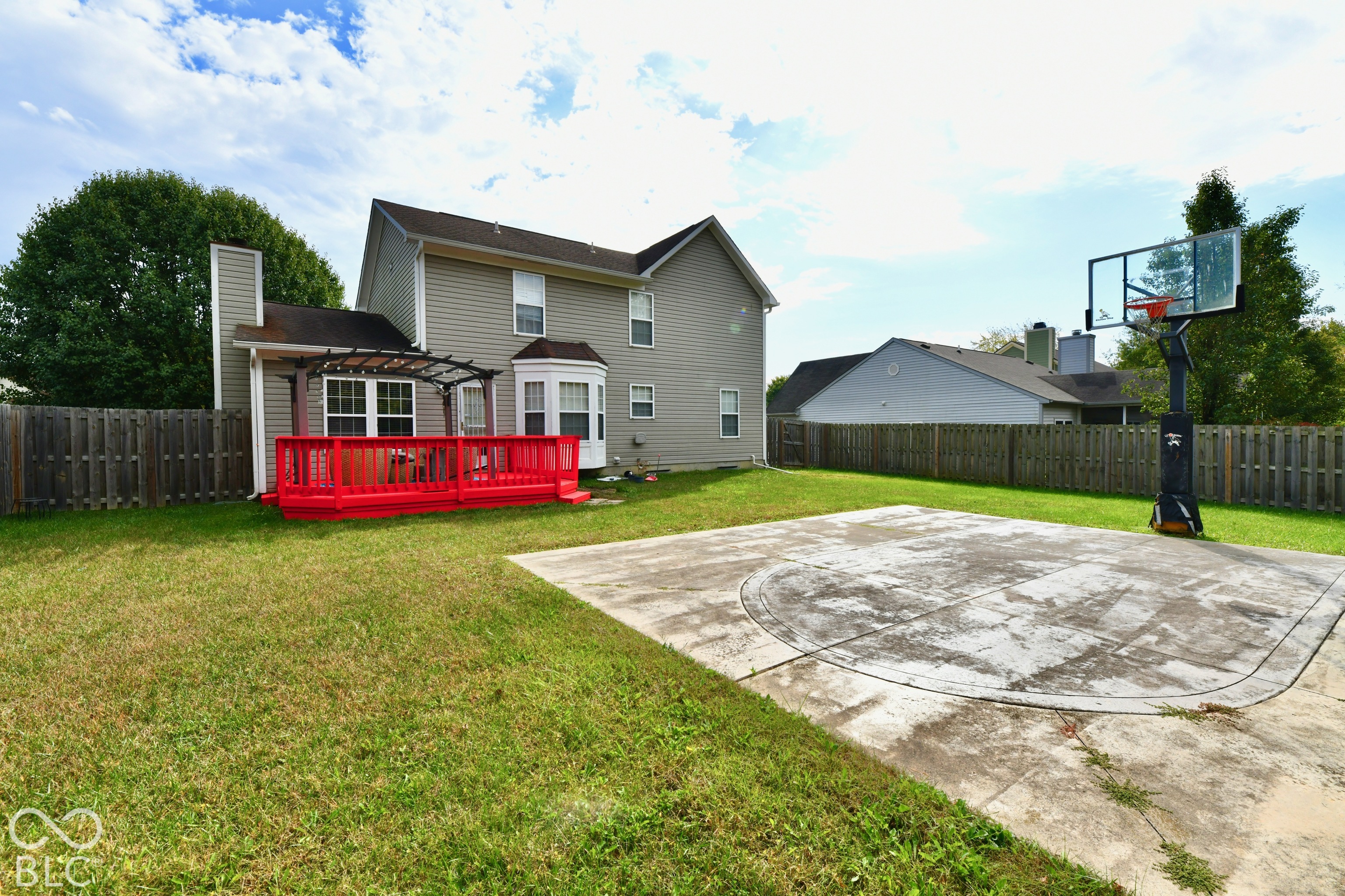 Rear exterior featuring a wood deck with an attached pergola, a paved concrete sports court, and perimeter wood fencing