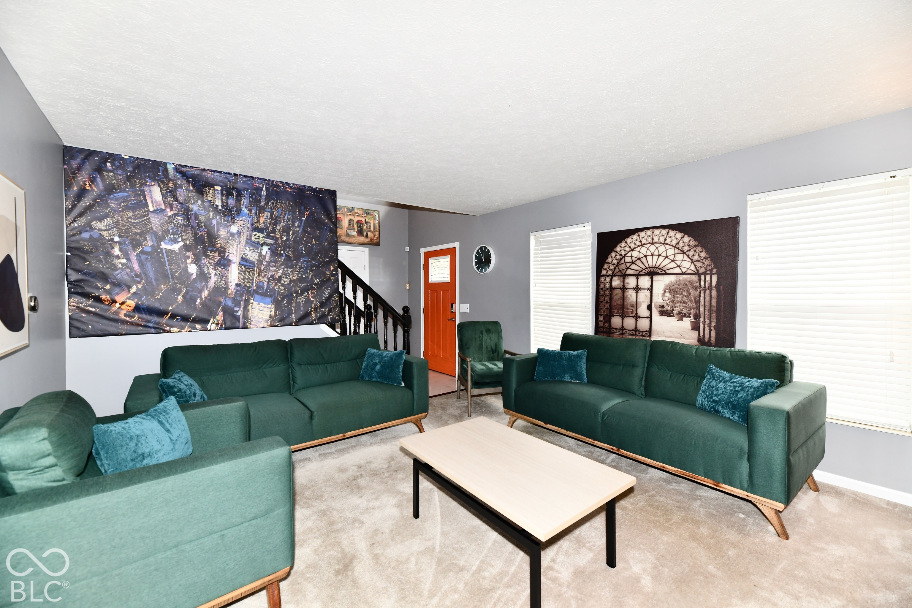 Living area featuring neutral carpeting, grey wall paint, horizontal window blinds, and a staircase with a dark wood banister