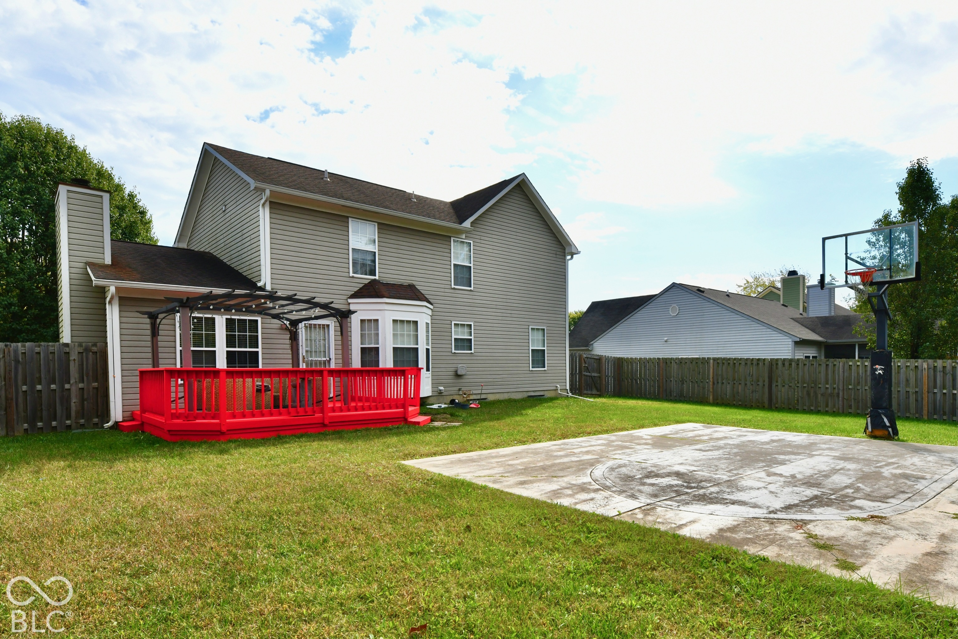 Fenced backyard featuring an elevated wood deck with a pergola, a paved recreation area, and a basketball goal