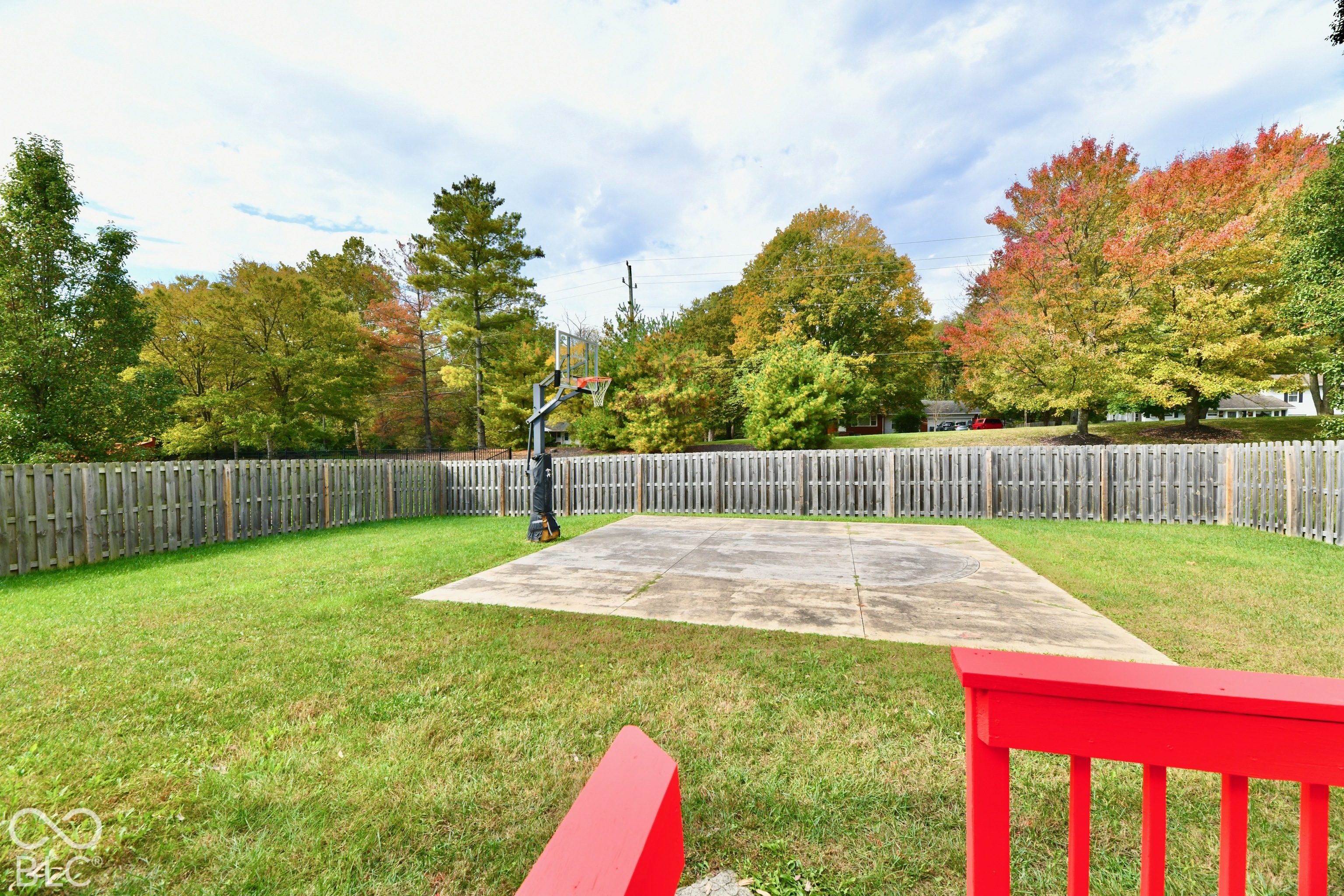 Backyard featuring a concrete court, attached basketball hoop, wood-plank fencing, and mature trees