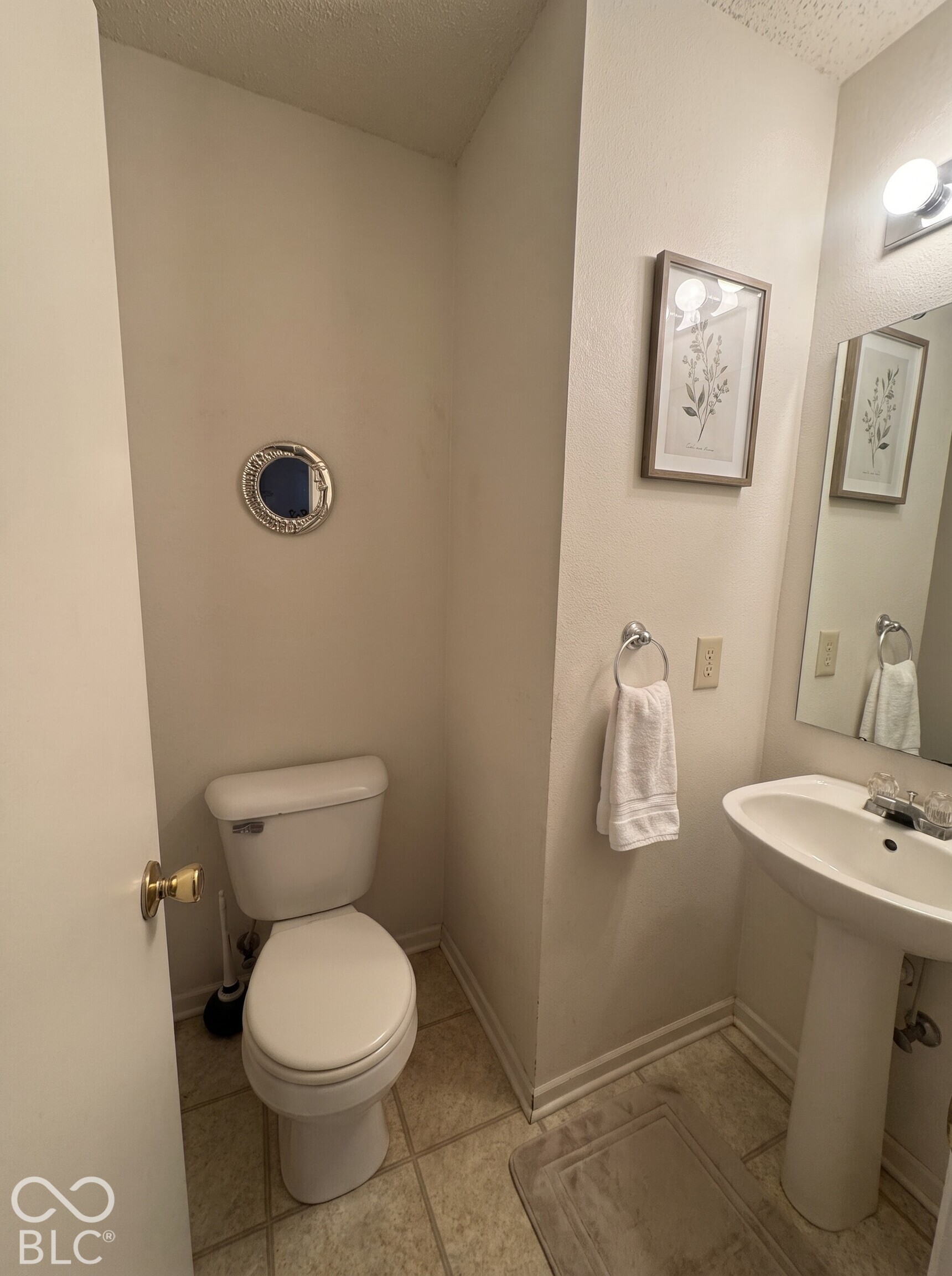 Virtually Altered - Powder room featuring a pedestal sink with chrome faucet, porcelain toilet, tiled flooring, and wall-mounted vanity lighting