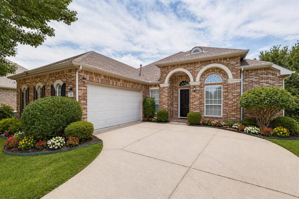 Brick exterior with arched window details, a curved concrete driveway, integrated two-car garage, and established landscaping with colorful plantings