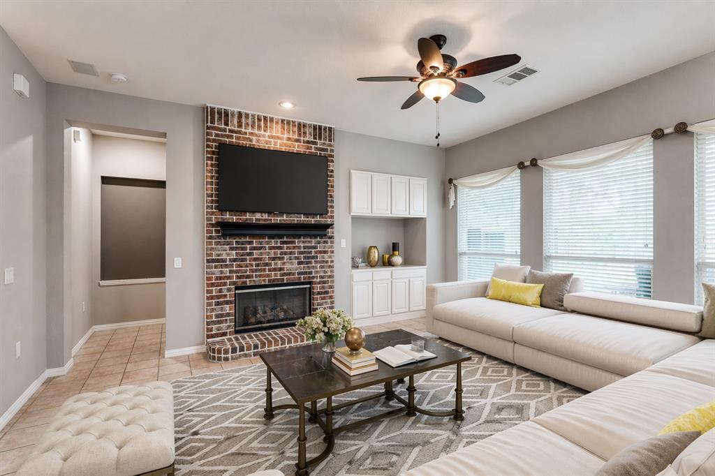 Living area featuring a brick fireplace, built-in cabinetry, multiple windows with blinds, a ceiling fan with integrated lighting, and tile flooring - Virtually Staged