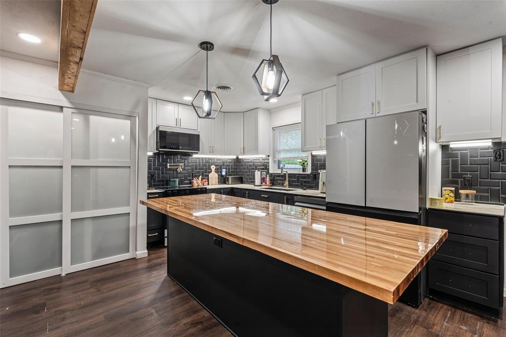 The kitchen features a large island with a wooden countertop, pendant lighting, hardwood flooring, and white upper cabinetry The kitchen features a large island with a wooden countertop, pendant lighting, hardwood flooring, and white upper cabinetry
