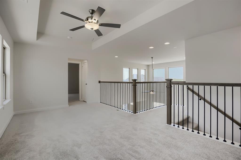 Carpeted loft area featuring a tray ceiling with a modern ceiling fan, large windows, and a dark wood and iron spindle railing overlooking an open space with additional windows