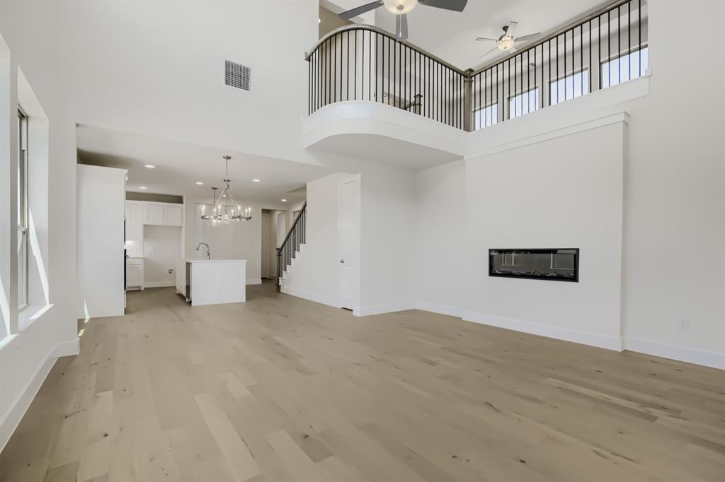 Open-concept interior featuring light wood-finish flooring, a contemporary fireplace, a kitchen island with a sink and chandelier, a staircase with dark railings, and a second-story balcony with dark balusters