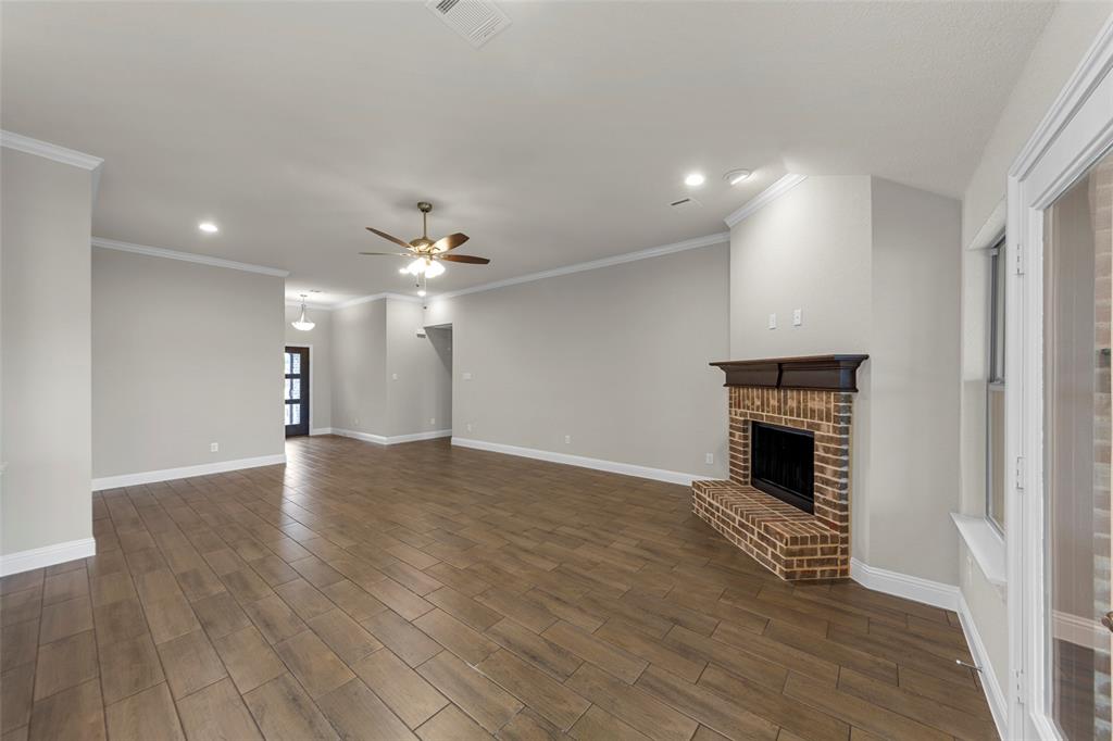Expansive open-concept living area featuring wood-finish flooring, a brick fireplace with a dark wood mantel, recessed lighting, and crown molding