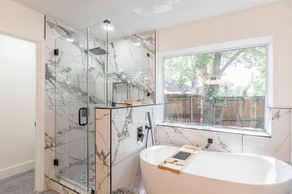 Spacious bathroom featuring a freestanding soaking tub, a large glass-enclosed shower with rain head, marbled tile surround, and expansive window