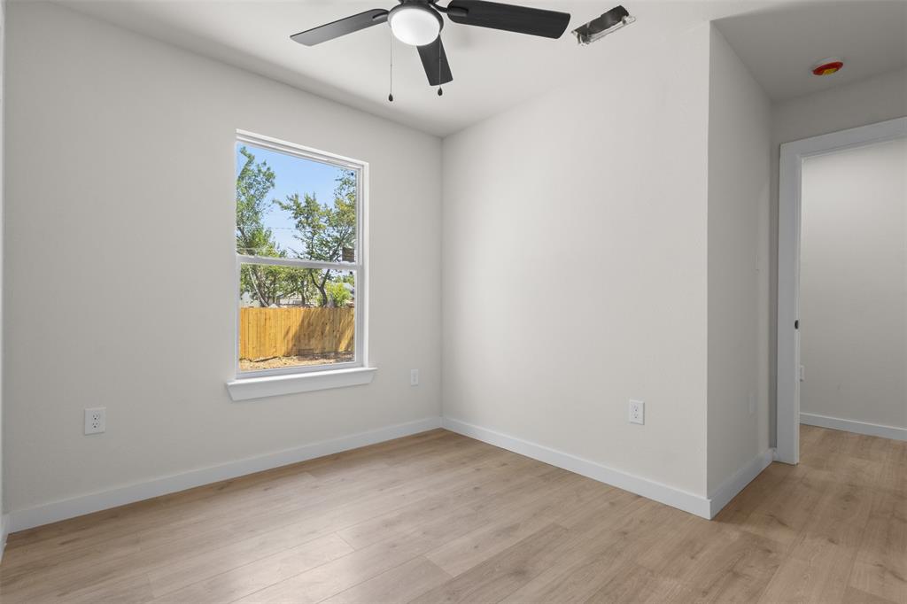 Bright, light-filled bedroom showcasing wood-finish flooring, a large double-hung window with natural landscaping views, ceiling fan with integrated lighting, and clean white walls with electrical outlets