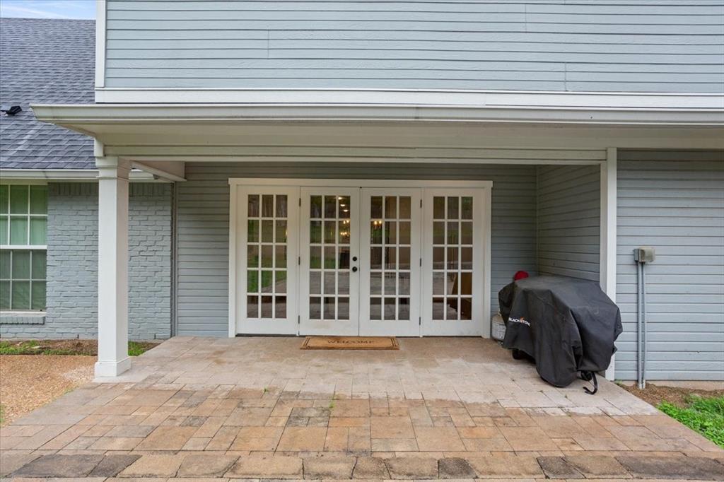 Paved patio featuring a covered overhang with white support columns, dual French doors with grid-patterned glass, and light blue siding