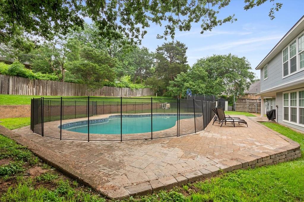 Backyard swimming pool with a black mesh safety fence, surrounded by a paver patio and mature trees
