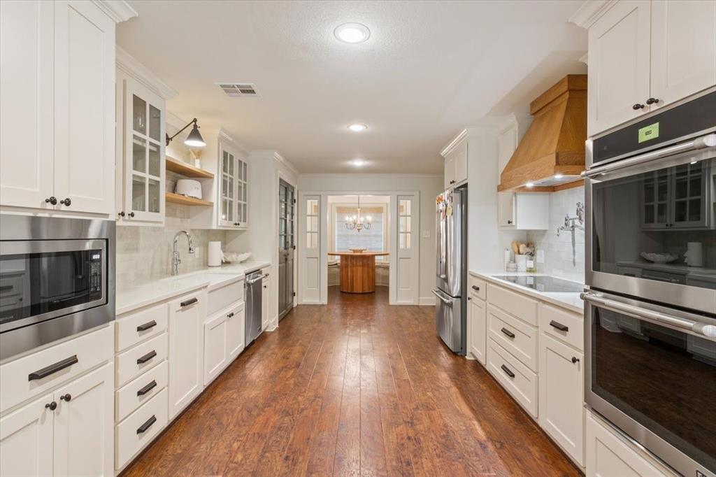 Expansive kitchen featuring wood-finish flooring, white cabinetry with dark hardware, stainless steel appliances, and a wood range hood Expansive kitchen featuring wood-finish flooring, white cabinetry with dark hardware, stainless steel appliances, and a wood range hood