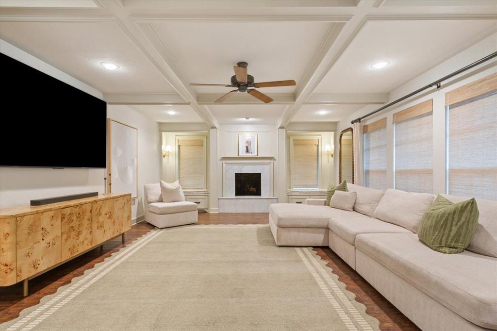 Living area featuring hardwood flooring, a coffered ceiling with recessed lighting and a ceiling fan, a fireplace with a stone surround and mantel, and multiple windows with roller shades Living area featuring hardwood flooring, a coffered ceiling with recessed lighting and a ceiling fan, a fireplace with a stone surround and mantel, and multiple windows with roller shades
