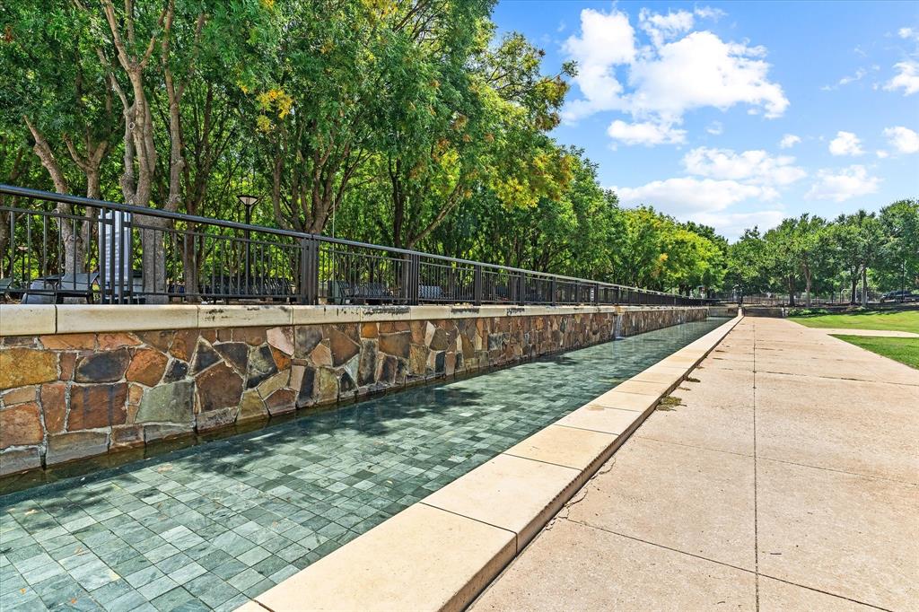 Linear water feature with a stone veneer wall and dark metal railing, bordered by concrete paving and mature trees