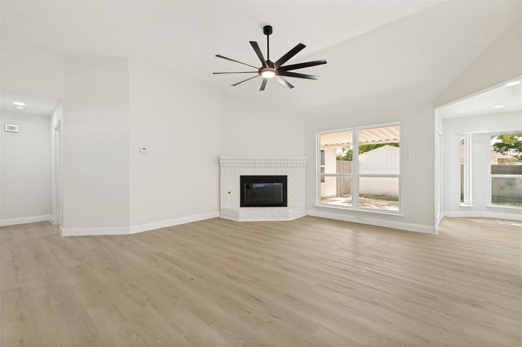Spacious living area featuring vaulted ceilings, wood-finish flooring, a white brick fireplace, a modern ceiling fan, and multiple large windows Spacious living area featuring vaulted ceilings, wood-finish flooring, a white brick fireplace, a modern ceiling fan, and multiple large windows