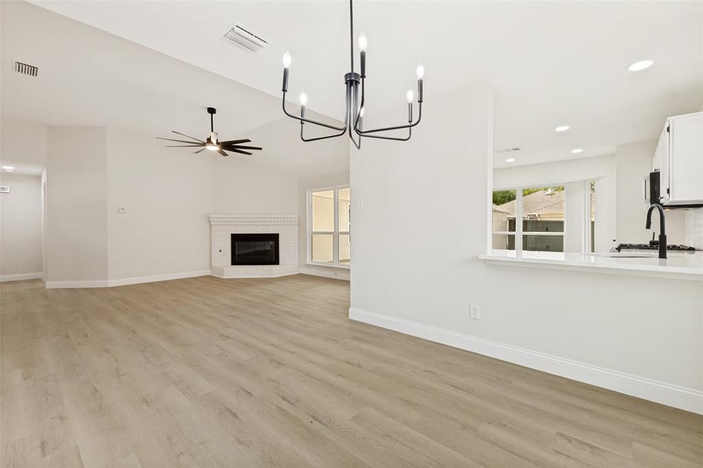Open-concept living space featuring wood-finish flooring, a contemporary black chandelier, a corner fireplace with a white brick surround, vaulted ceilings, and a kitchen pass-through with a white countertop
