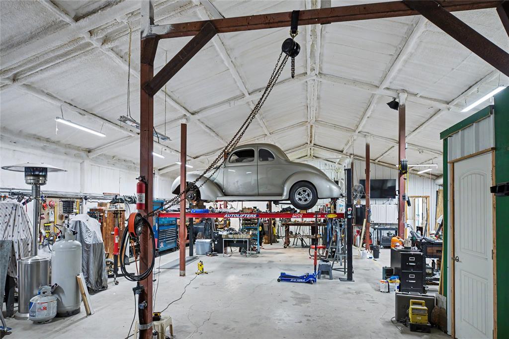Spacious garage workshop featuring a vehicle lift, vaulted ceiling with exposed beams, fluorescent lighting, and a fire extinguisher