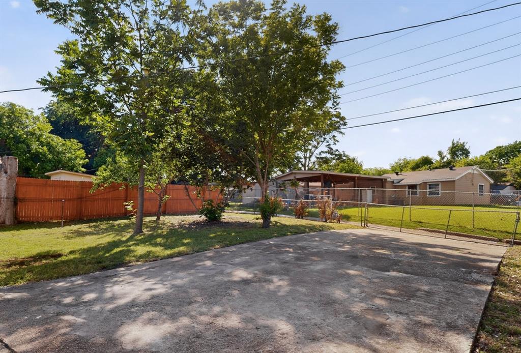 Gated concrete driveway with a chain-link fence, mature trees, and a wooden privacy fence bordering a grass lawn