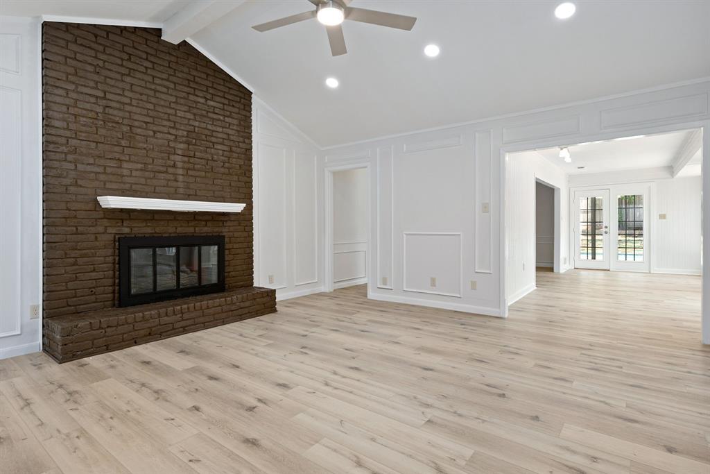 Spacious living area featuring a prominent brick fireplace with a white mantel, vaulted ceilings with exposed beams, recessed lighting, and wood-finish flooring Spacious living area featuring a prominent brick fireplace with a white mantel, vaulted ceilings with exposed beams, recessed lighting, and wood-finish flooring