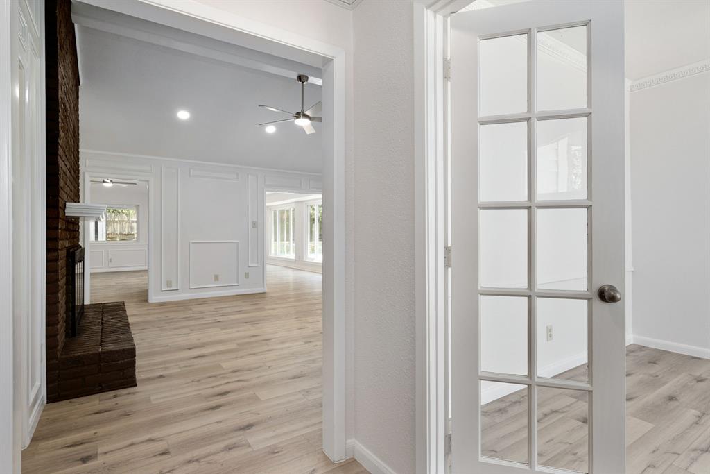 French doors open to an expansive living area featuring wood-finish flooring, a brick fireplace, a vaulted ceiling, recessed lighting, and decorative wall paneling French doors open to an expansive living area featuring wood-finish flooring, a brick fireplace, a vaulted ceiling, recessed lighting, and decorative wall paneling
