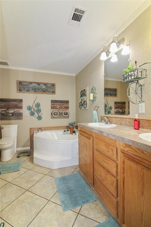Bathroom featuring a corner soaking tub with deck-mounted fixtures, dual vanity with wood-finish cabinetry, ceramic tile flooring, and a standard toilet