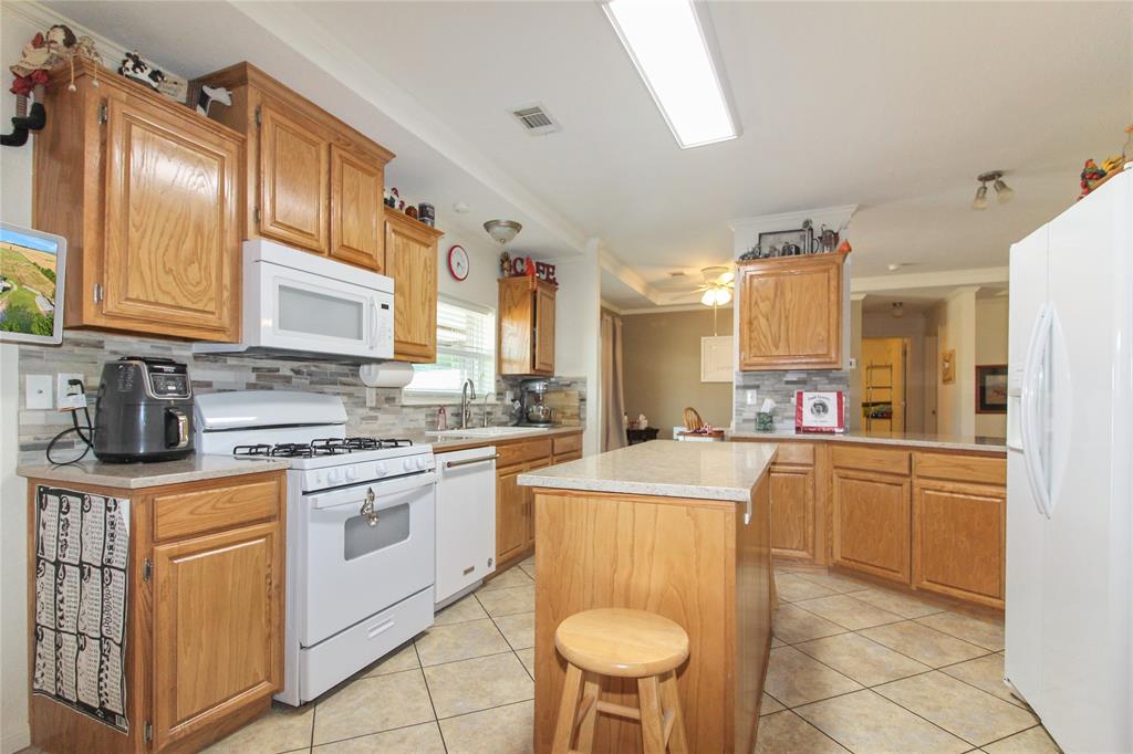 Kitchen featuring light wood-finish cabinetry, light-toned stone-finish countertops, a tile backsplash, white appliances, and tile flooring