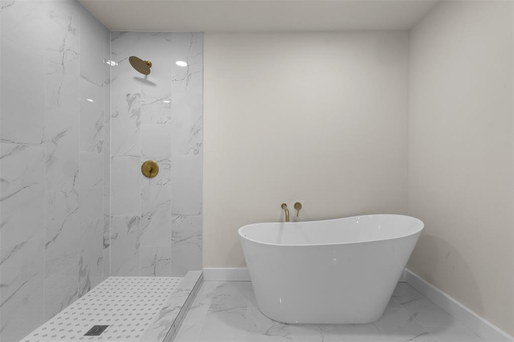 Freestanding soaking tub with wall-mounted brass fixtures, marble-look tile flooring, and a walk-in shower featuring matching marble-look wall tiles and brass fixtures