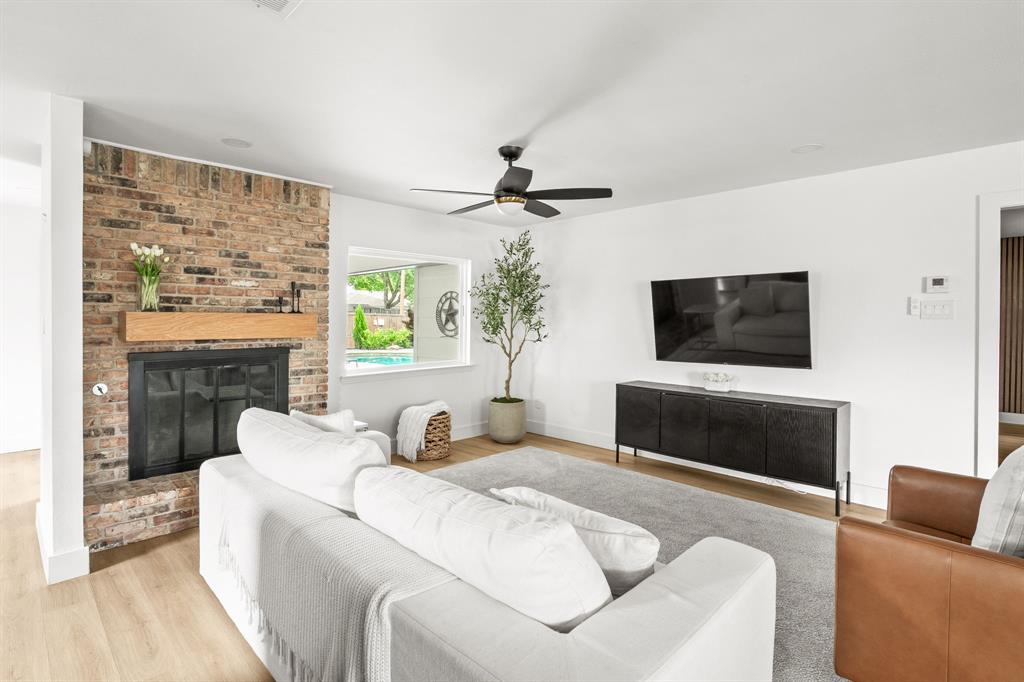 Living area showcasing a brick fireplace with a wood mantle, large window overlooking a pool, ceiling fan, and wood-finish flooring Living area showcasing a brick fireplace with a wood mantle, large window overlooking a pool, ceiling fan, and wood-finish flooring
