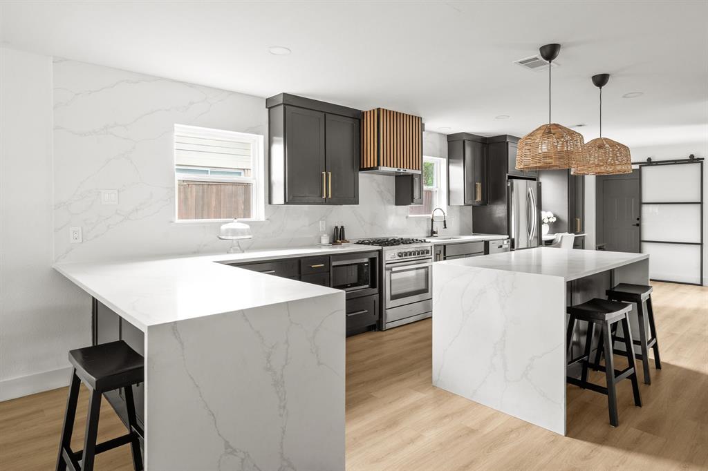Contemporary kitchen featuring an expansive waterfall island, wood-finish flooring, dark cabinetry with brass hardware, a full-height marble-look backsplash, and recessed lighting
