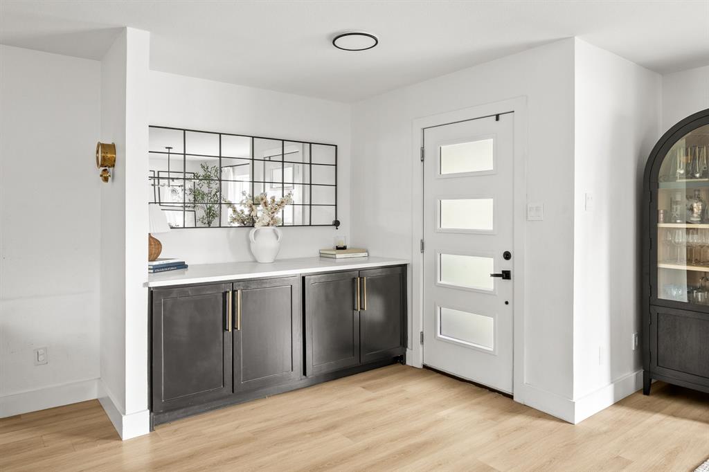 Entryway featuring a white door with frosted glass panels, a built-in dark cabinetry with gold-tone hardware and white countertop, and light wood-finish flooring Entryway featuring a white door with frosted glass panels, a built-in dark cabinetry with gold-tone hardware and white countertop, and light wood-finish flooring