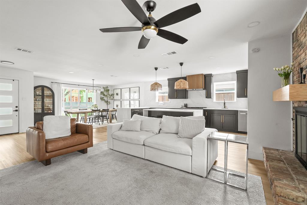 Open-concept living area featuring light wood-finish flooring, a contemporary ceiling fan, a brick fireplace with a wood mantle, a modern kitchen with dark cabinetry, and white countertops Open-concept living area featuring light wood-finish flooring, a contemporary ceiling fan, a brick fireplace with a wood mantle, a modern kitchen with dark cabinetry, and white countertops