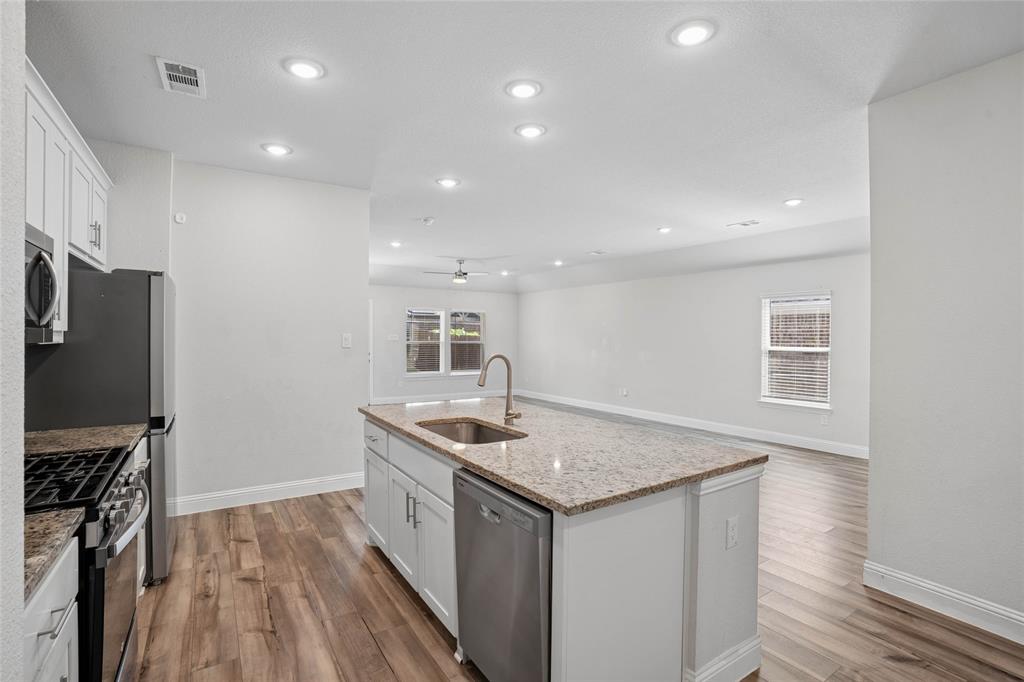 Open-concept kitchen featuring an expansive center island with an undermount sink, granite-style countertops, white cabinetry, and wood-finish flooring Open-concept kitchen featuring an expansive center island with an undermount sink, granite-style countertops, white cabinetry, and wood-finish flooring