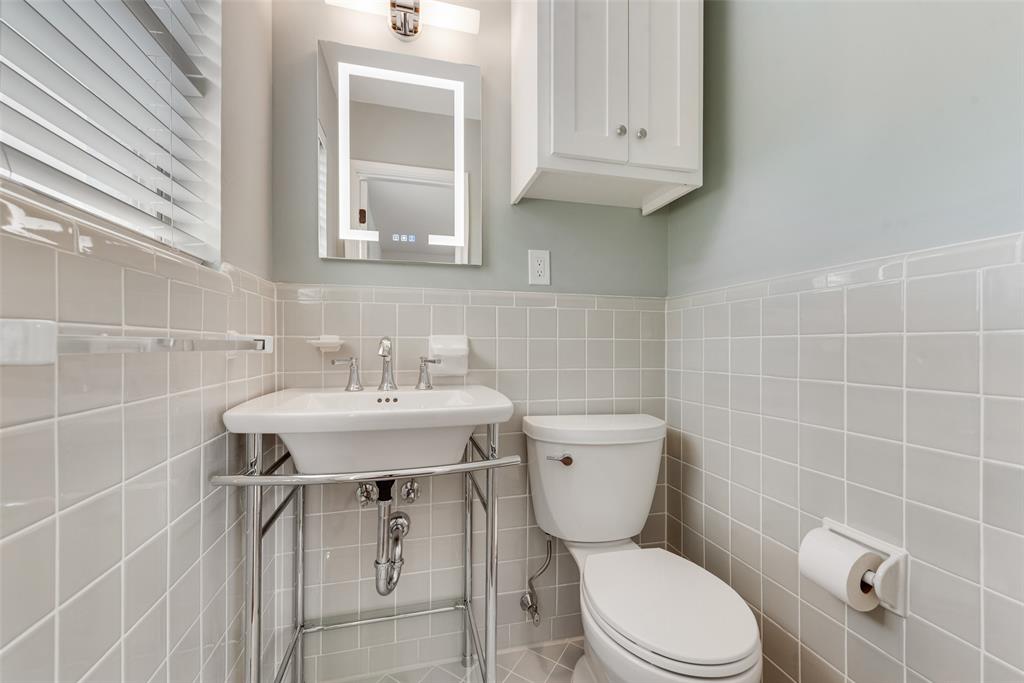Bathroom featuring light grey square wall tiling, a pedestal sink with chrome stand, a wall-mounted lighted vanity mirror, and a white storage cabinet