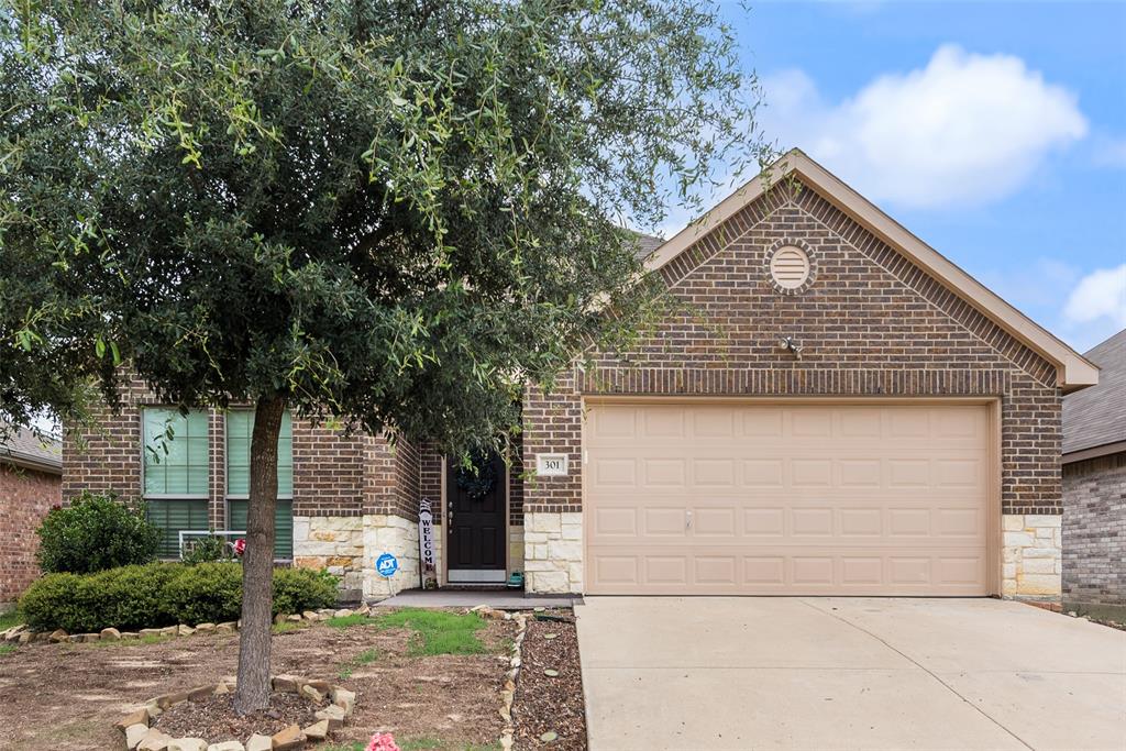 Brick and stone facade residence featuring a two-car garage, mature front yard tree, concrete driveway, and front entryway with a dark-paneled door