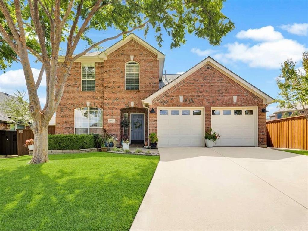 Brick facade residence featuring a two-car garage with panel windows, a concrete driveway, a manicured lawn, and mature trees