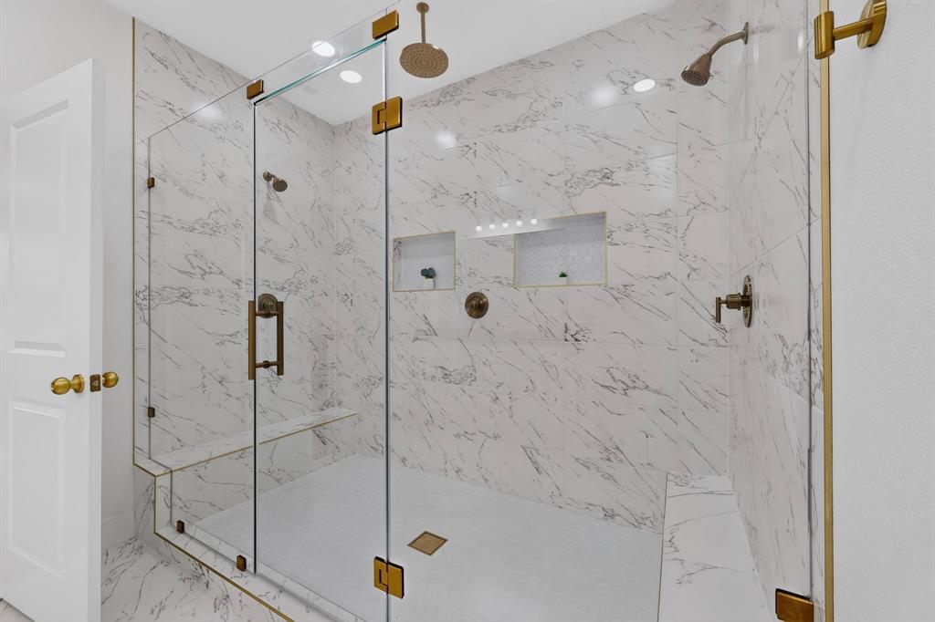 Spacious glass-enclosed shower featuring large format marble-look tile, dual shower heads, integrated bench seating, recessed niches, and brass-finish hardware