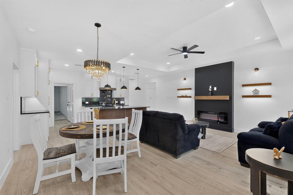 Open-concept living space featuring wood-finish flooring throughout, a modern fireplace with a dark accent wall and wood mantel, and tray ceilings
