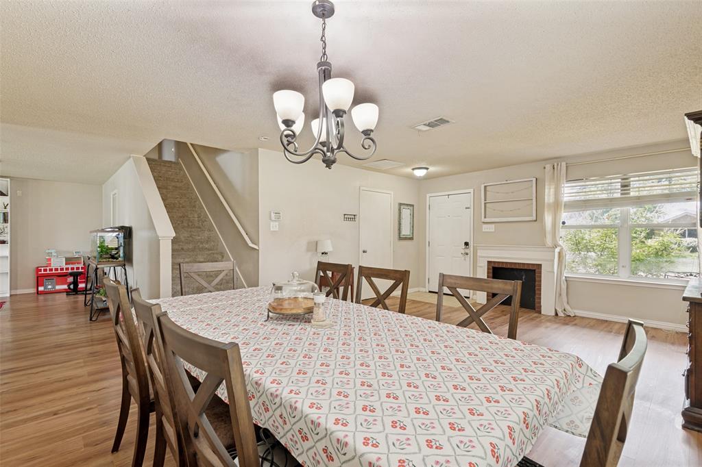 Open-concept living area featuring wood-finish flooring, a painted brick fireplace with a white mantel, and a large window with white blinds