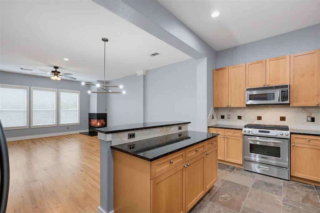 Kitchen and living area featuring a two-tiered island with a black countertop, natural wood-finish cabinetry, stainless steel appliances, and a tiled backsplash