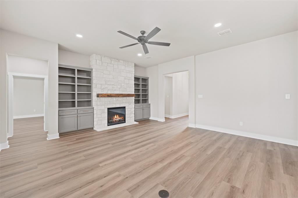 Spacious room featuring wood-finish flooring, a stone-clad fireplace with a wood mantel, and built-in shelving with lower cabinetry Spacious room featuring wood-finish flooring, a stone-clad fireplace with a wood mantel, and built-in shelving with lower cabinetry