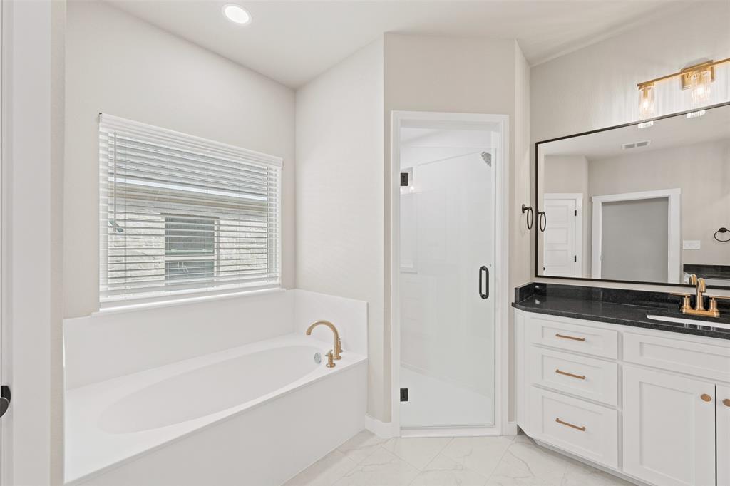 Bathroom featuring a built-in soaking tub with brass fixtures, a walk-in shower with a frameless glass door, a double vanity with a dark countertop and gold-toned hardware, and a large wall-mounted mirror