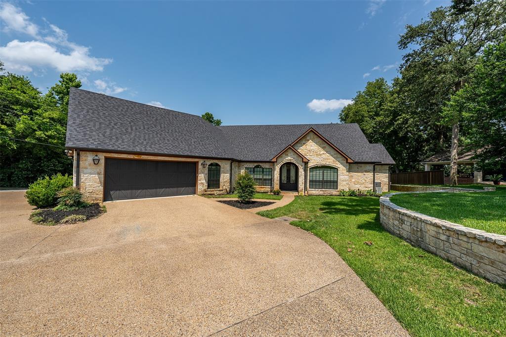 Stone facade residence featuring an expansive driveway, a two-car garage, and a dark shingle roof
