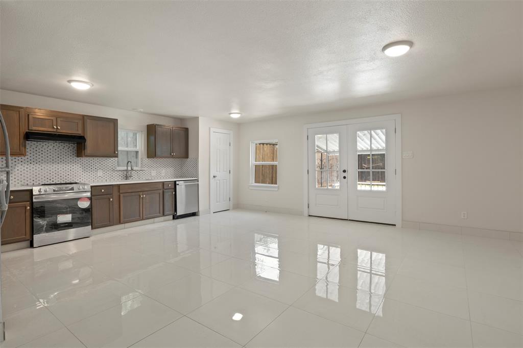 Open-concept kitchen and living area featuring white tile flooring, brown cabinetry, stainless steel appliances, and a mosaic tile backsplash
