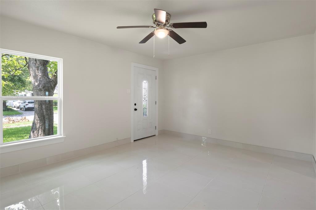 Spacious room featuring light-toned flooring, a ceiling fan with integrated lighting, a white entry door with decorative glass, and a large window providing outdoor illumination
