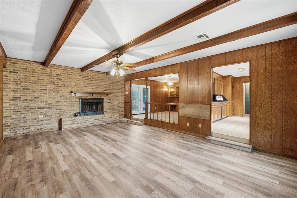 Spacious living area featuring wood-finish flooring, a brick fireplace with a mantel, exposed wood ceiling beams, and a ceiling fan