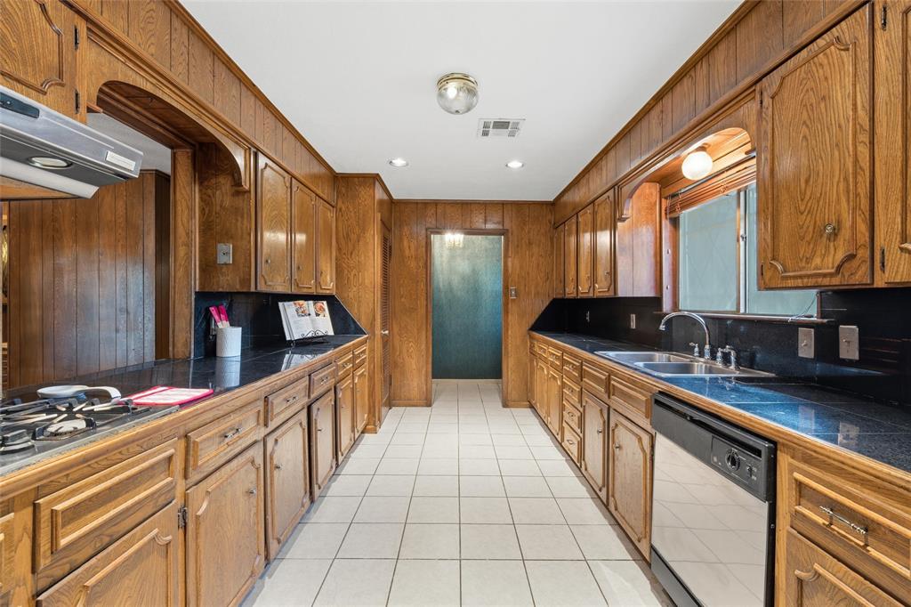 Galley kitchen featuring extensive wood cabinetry, dark countertops, white tile flooring, stainless steel gas range, and a stainless steel dishwasher