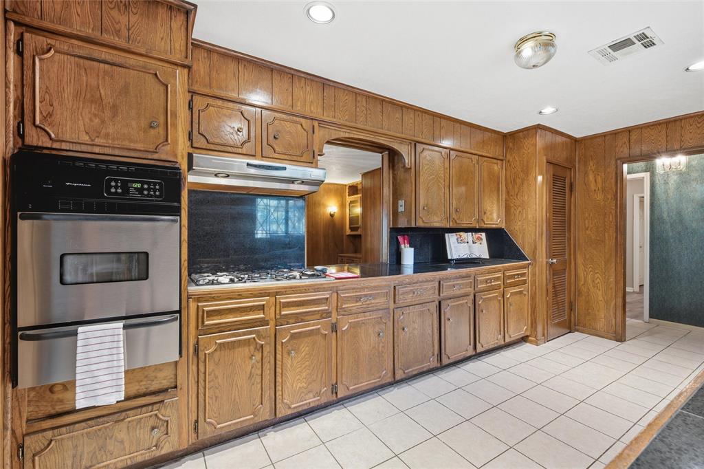 Kitchen featuring wood-paneled walls, built-in double oven, gas cooktop with exhaust hood, dark countertops, and white square tile flooring