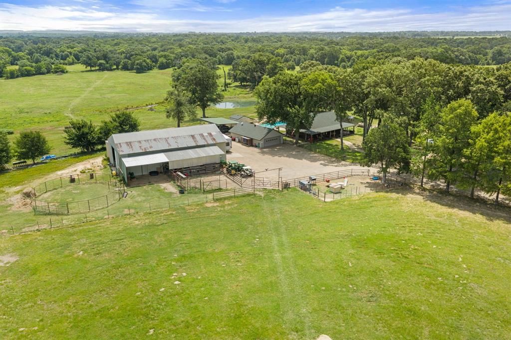 Expansive property featuring a large barn with an attached lean-to, multiple fenced enclosures, a separate building with a green roof, and a visible pond surrounded by mature trees