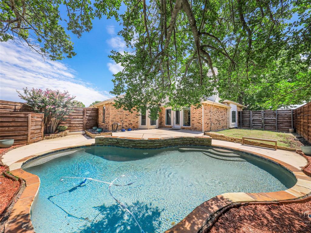 Backyard oasis featuring a curvilinear swimming pool with integrated steps, stone coping, and a surrounding concrete patio.  Note the pool equipment is located at the back of the lot near the driveway and garage behind a board on board fence to protect and hide the equipment.