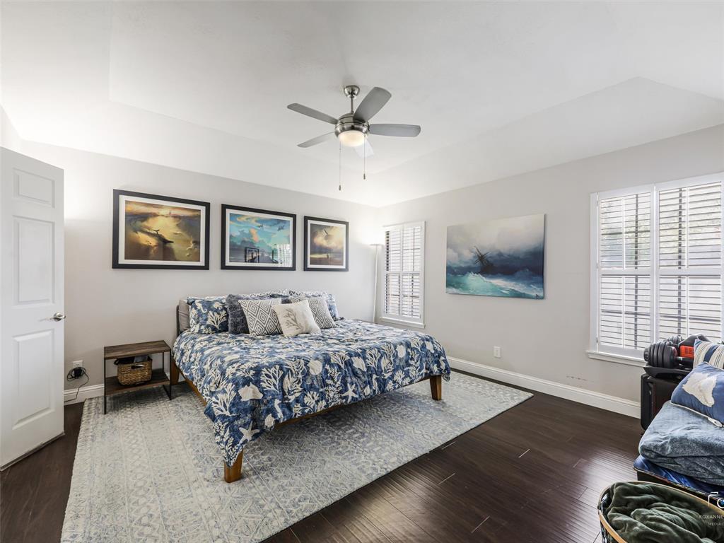 The same dark wood floors continue into the primary bedroom, along with a ceiling fan and windows with shutters.